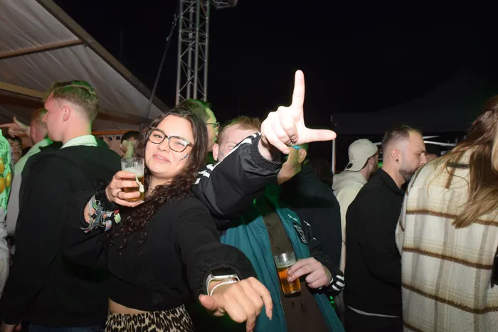 Eine junge Frau mit dunklen lockigen Haaren und Brille hält ein Bierglas hoch und zeigt mit der anderen Hand auf eine Person im Hintergrund. Um sie herum tanzen und feiern Menschen auf einer Party unter einem Zelt.