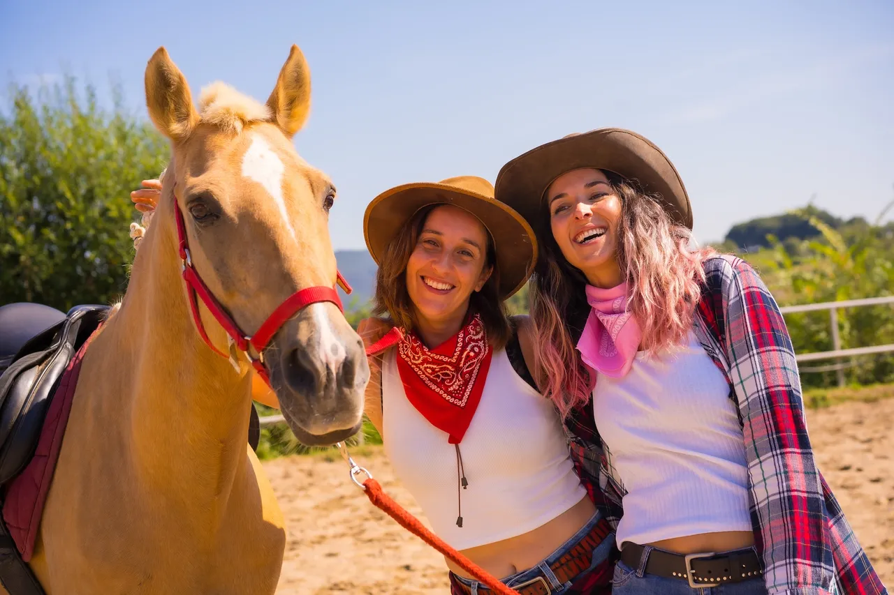 Zwei lächelnde Frauen mit Cowboyhüten stehen neben einem hellbraunen Pferd auf einem staubigen Feld. Beide tragen rote Halstücher und schauen in die Kamera.