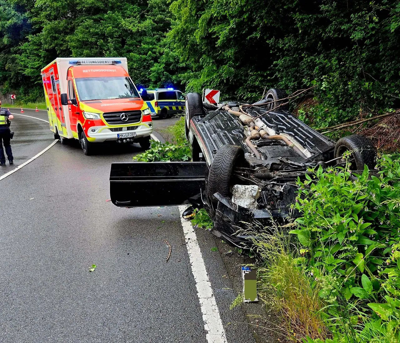 Umgestürztes Auto an einer Landstraße, neben ihm ein Rettungswagen und Einsatzkräfte. Im Hintergrund Bäume und ein Warnschild.