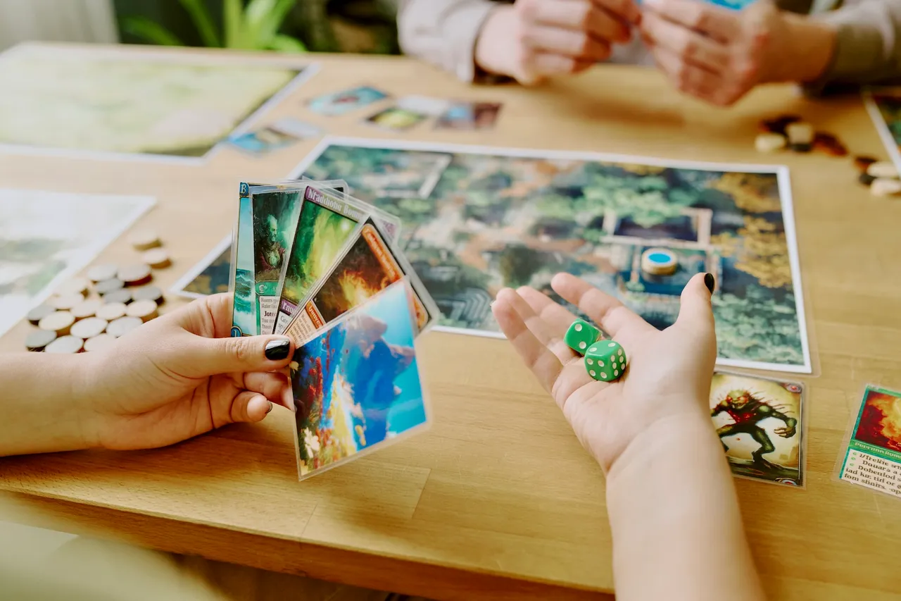 Zwei Personen spielen ein Brettspiel mit Karten und Würfeln auf einem Holztisch. Im Hintergrund sind weitere Spielmaterialien und Mitspieler zu sehen.