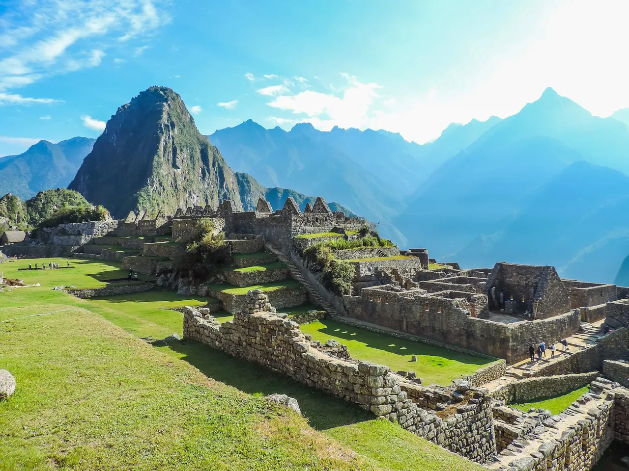 Steinruinen und grüne Terrassen der Inka-Stätte Machu Picchu vor einer Kulisse aus steilen Bergen unter blauem Himmel.
