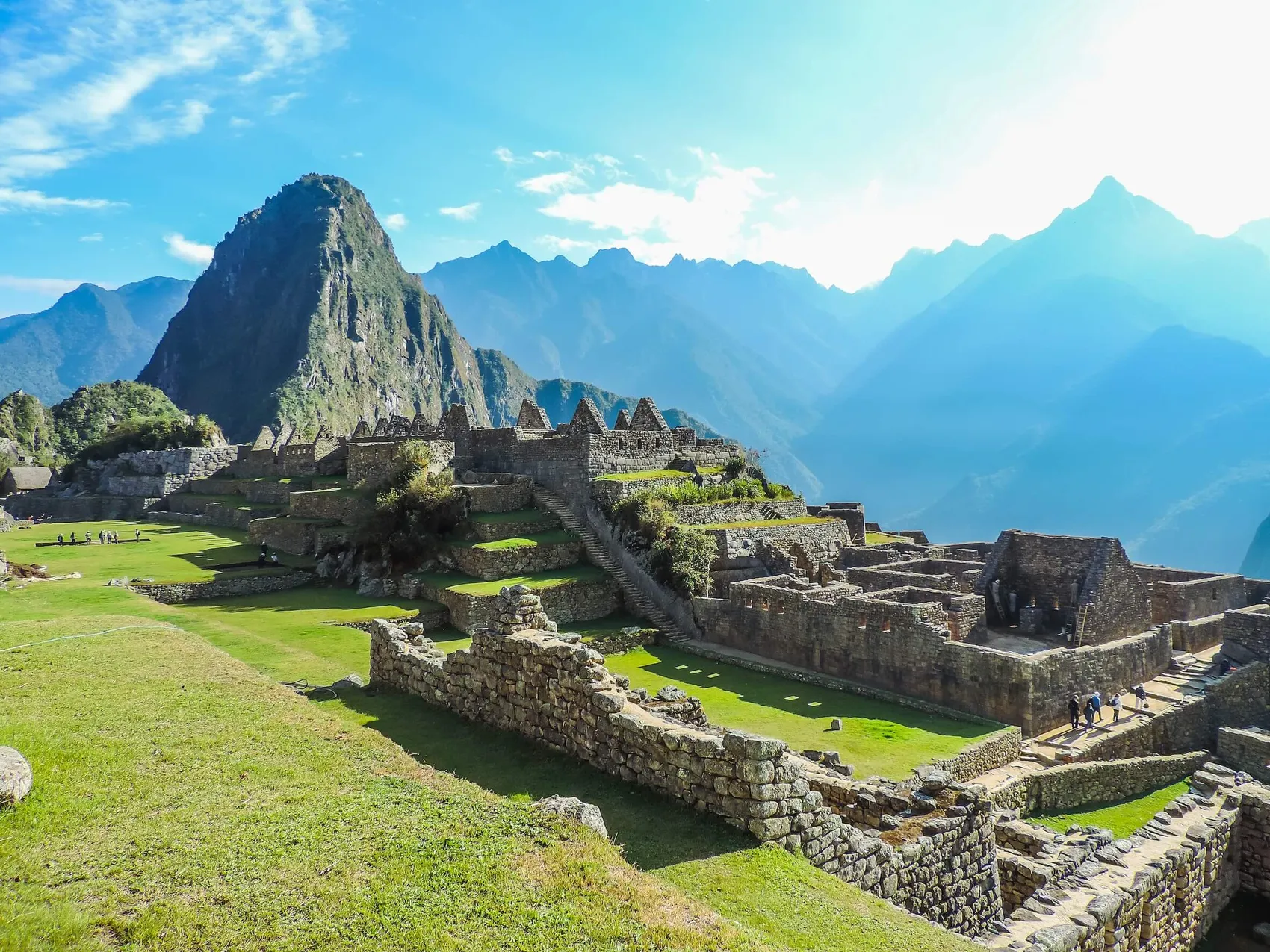 Steinruinen und grüne Terrassen der Inka-Stätte Machu Picchu vor einer Kulisse aus steilen Bergen unter blauem Himmel.