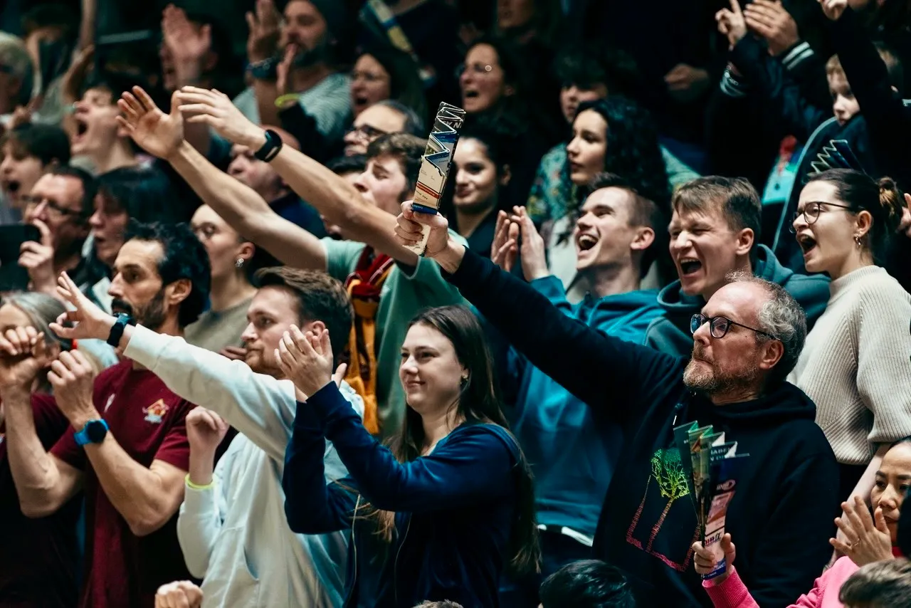 Jubelnde Fans mit erhobenen Händen in einer Halle, ein Mann hält eine Fahne hoch.