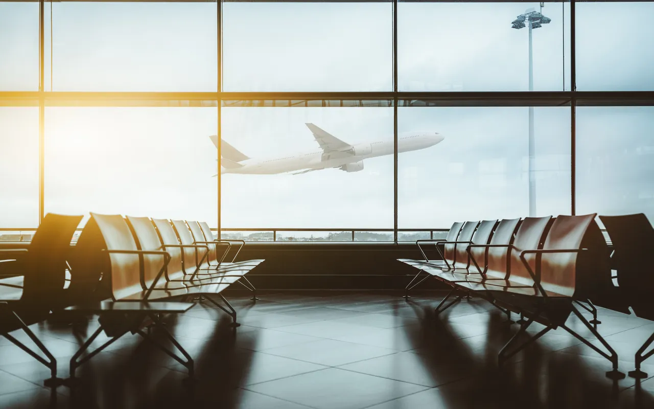 Ein Flugzeug startet vor einer großen Fensterfront in einer menschenleeren Flughafenhalle mit Reihen von Sitzbänken.