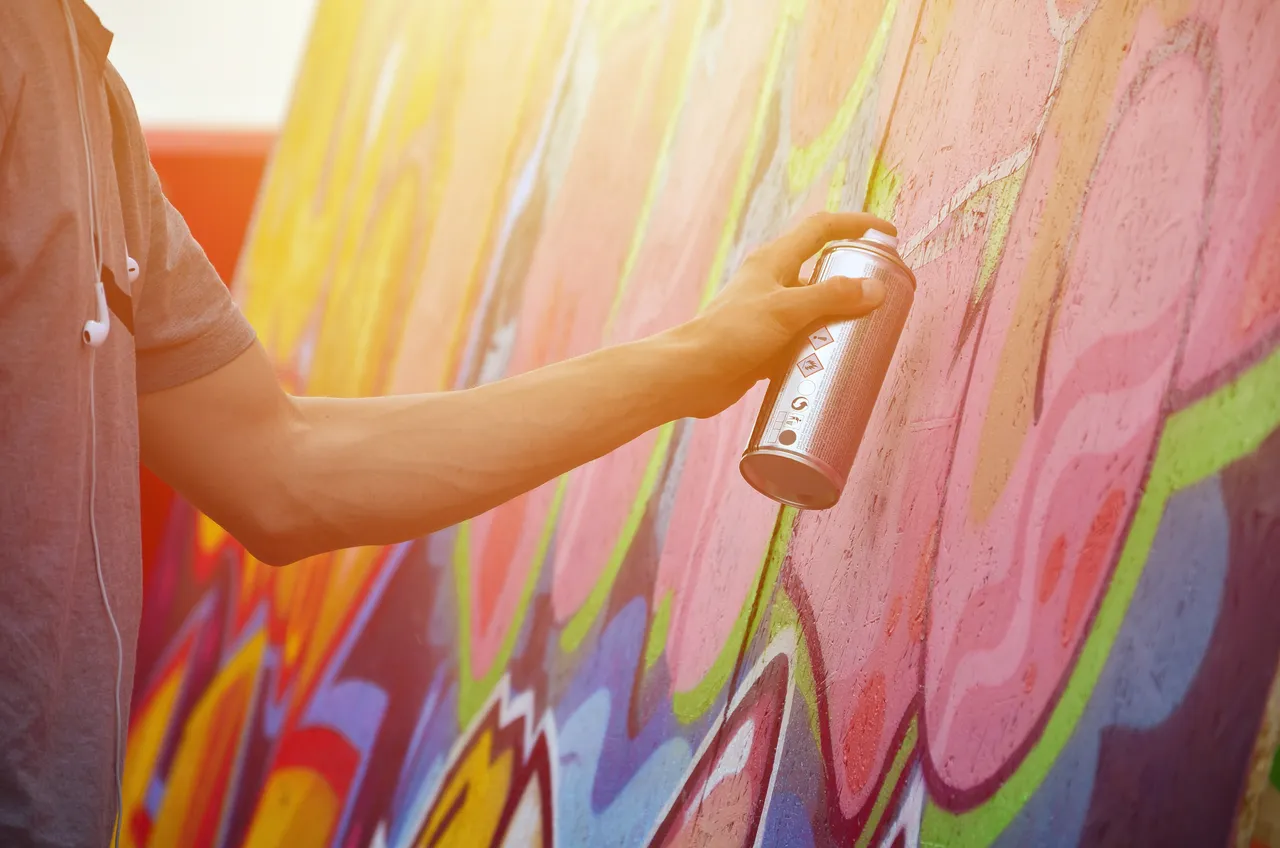 Eine Person sprüht mit einer Spraydose bunte Farben auf eine Mauer und gestaltet so ein Graffiti.