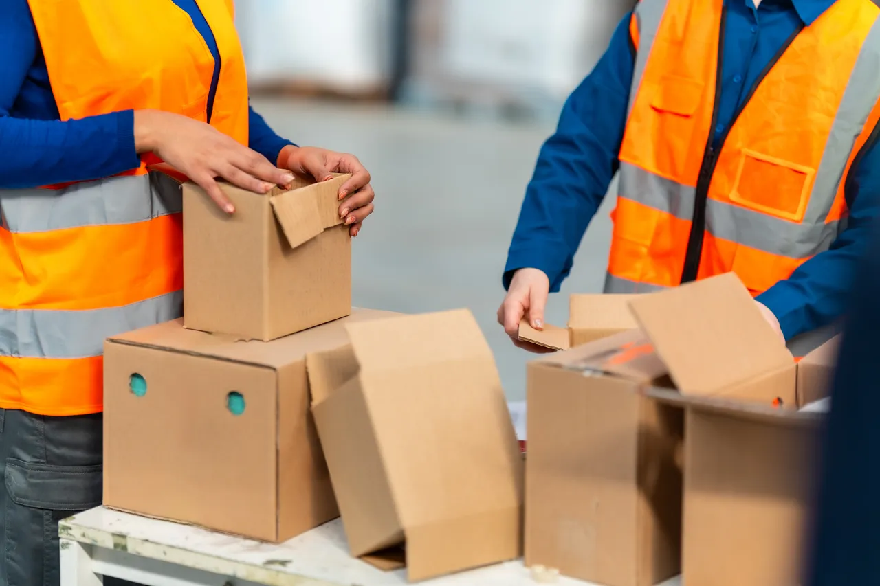 Mitarbeiter in orangefarbenen Warnwesten packen braune Kartons für den Versand in einem Lager. Die neuen Vorschriften betreffen auch Verpackungen und Kennzeichnungen.