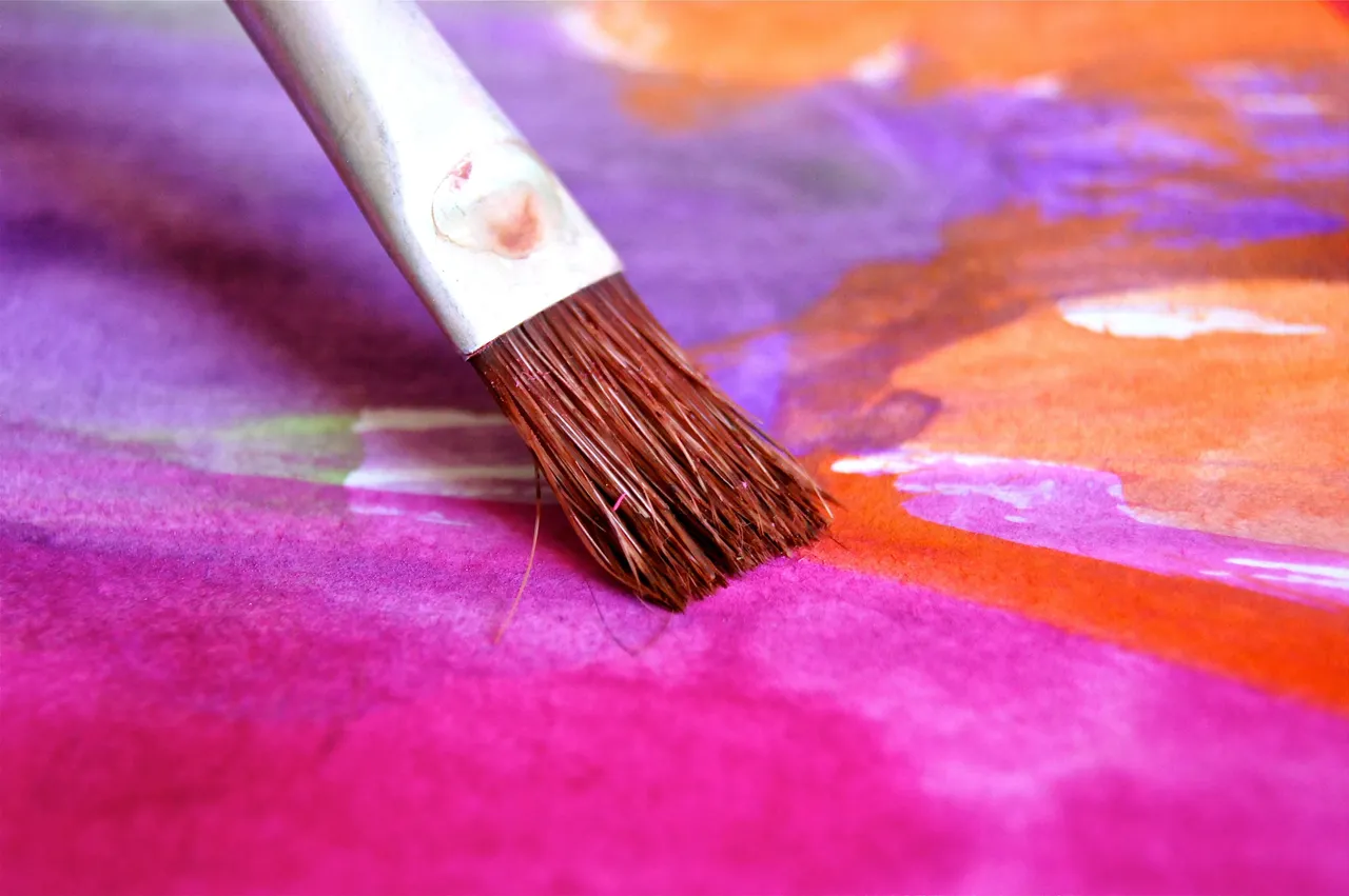 Nahaufnahme eines Pinsels auf einer farbigen Leinwand mit violetten, orangefarbenen und rosafarbenen Farbtönen.