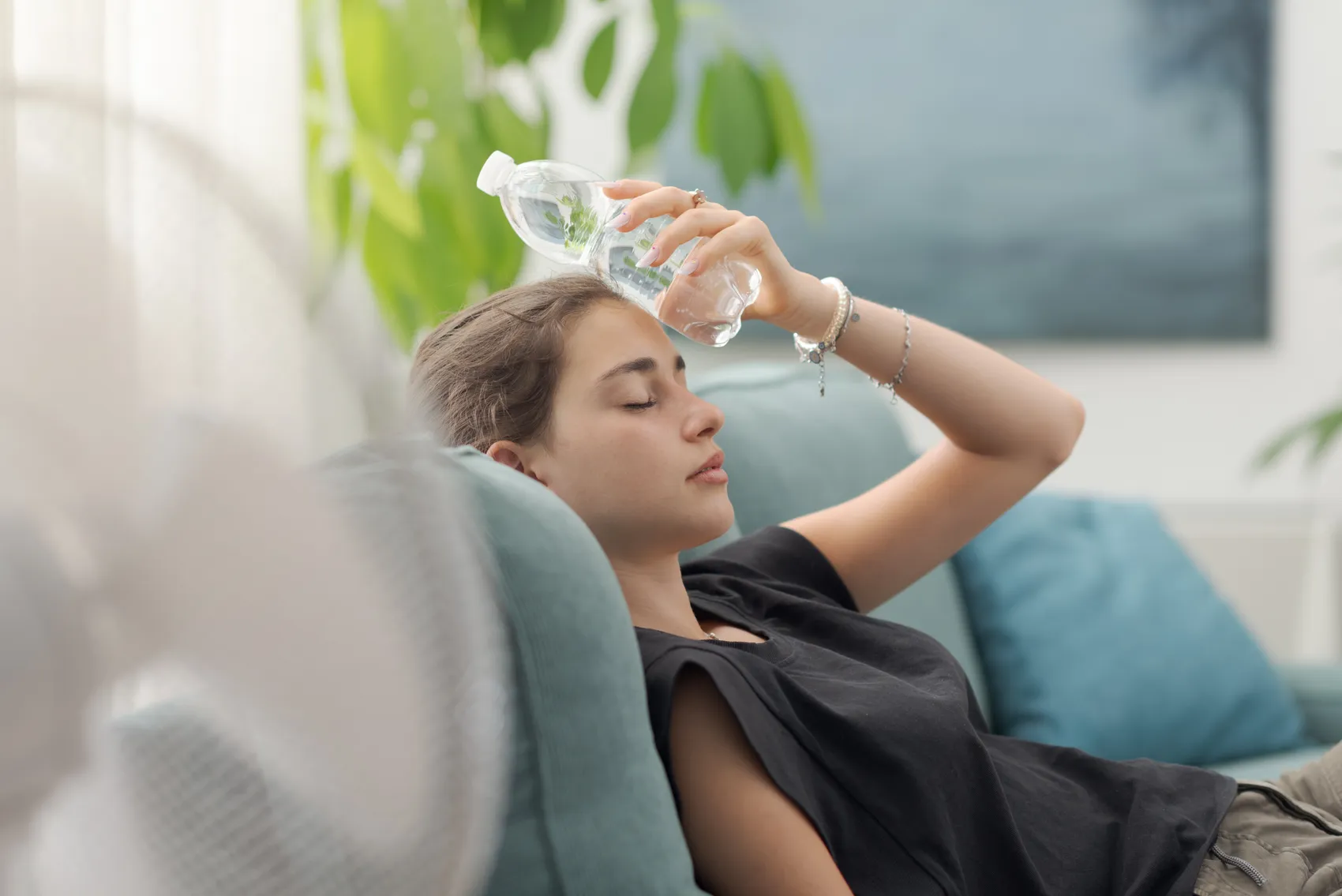Eine Frau liegt auf einer Couch und kühlt ihren Kopf mit einer Wasserflasche ab. Sie scheint sich unwohl zu fühlen, möglicherweise aufgrund von Hitze.