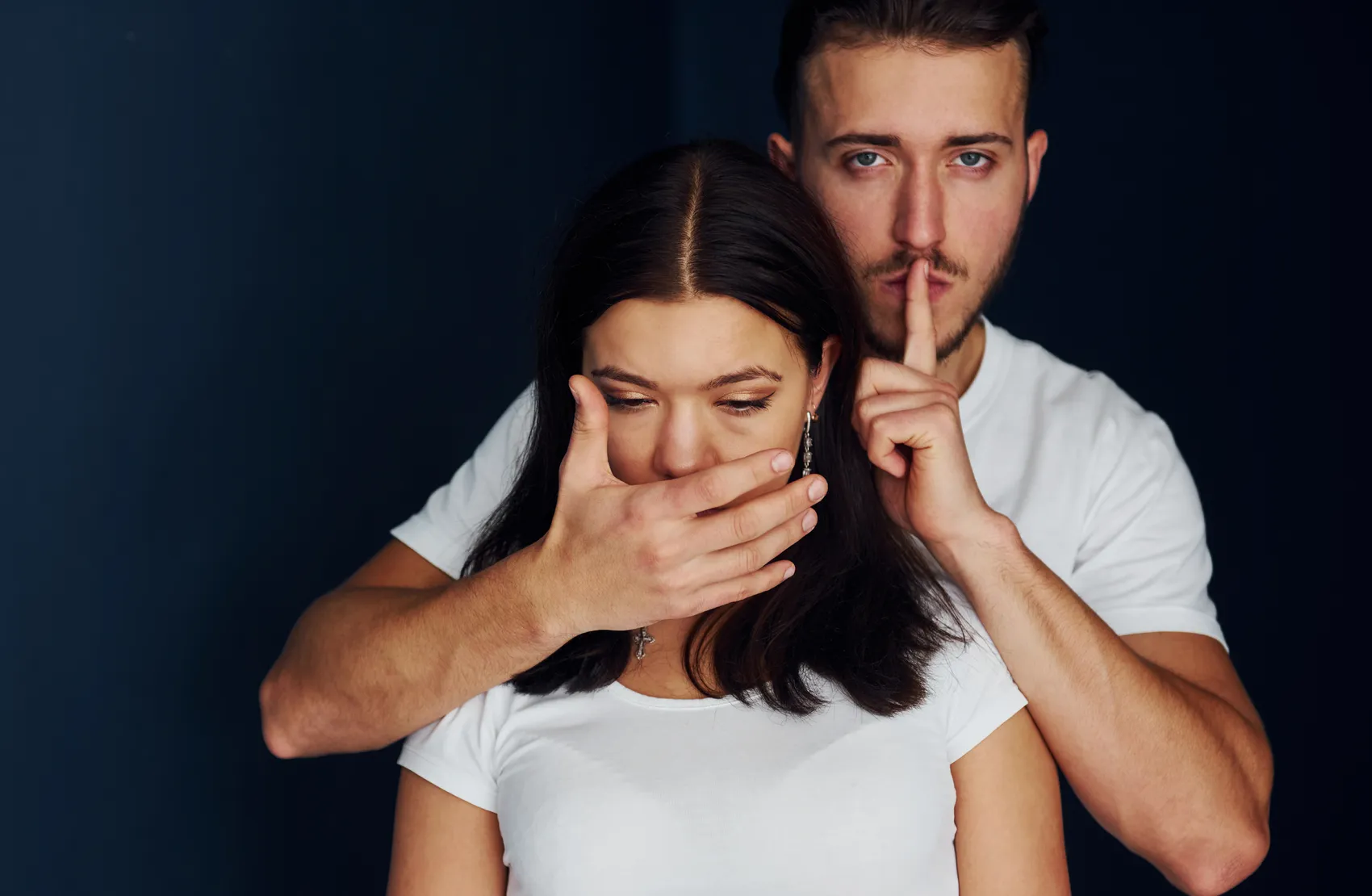 Ein Mann hält einer Frau die Hand vor den Mund und deutet mit dem Zeigefinger auf die Lippen, um Stille zu signalisieren. Das Bild steht symbolisch für sexualisierte Gewalt und deren Tabuisierung.