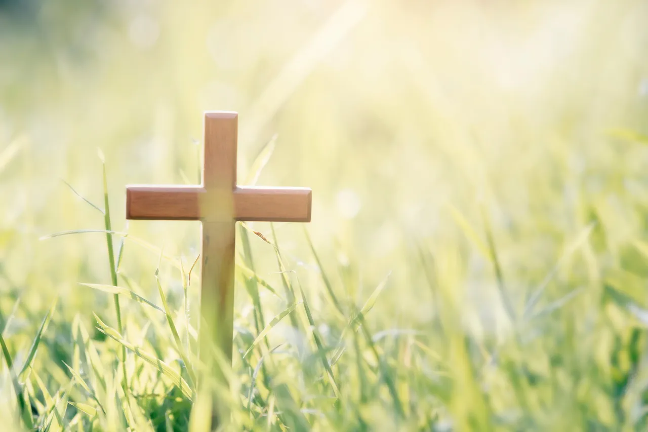 Holzkreuz steht im grünen Gras, im Hintergrund verschwommene Lichtstimmung.