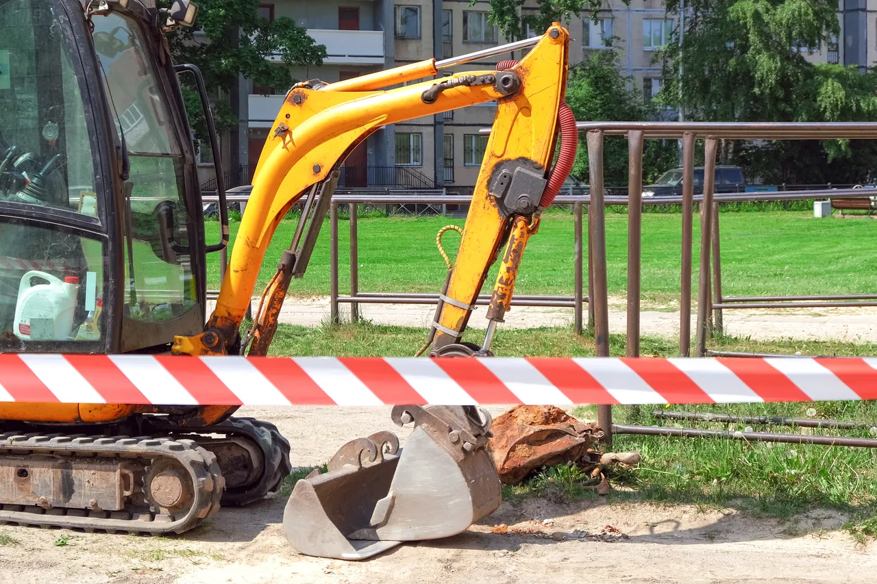 Gelber Bagger mit Schaufel vor einer Absperrung und einem grünen Park im Hintergrund.