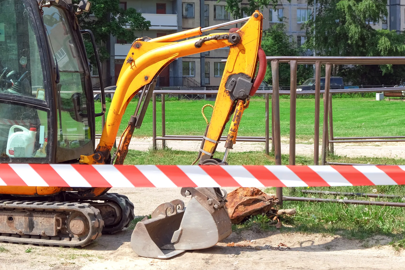 Gelber Bagger mit Schaufel vor einer Absperrung und einem grünen Park im Hintergrund.