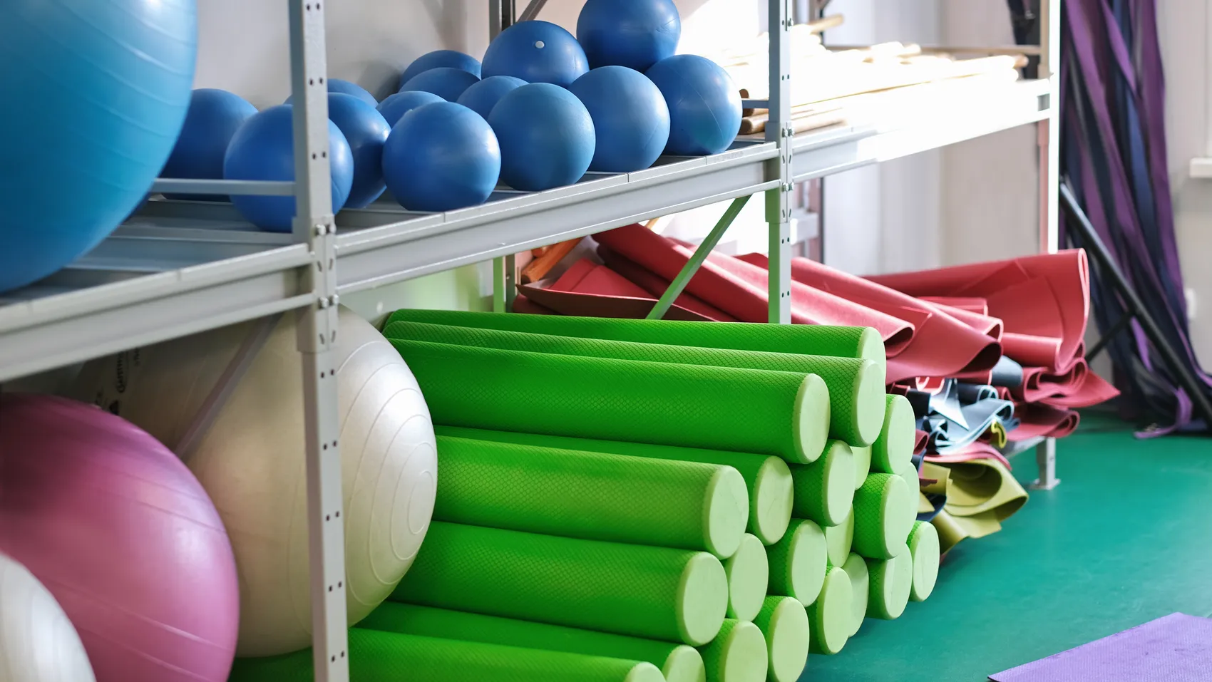 Mehrere grüne Yogamatten und blaue Gymnastikbälle stehen auf Regalen in einer Turnhalle bereit.