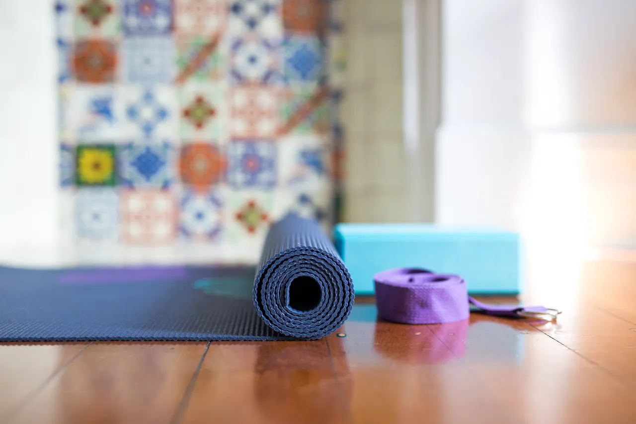 Gerollte blaue Yogamatte, ein blauer Block und ein lilafarbener Gurt liegen auf einem Holzboden vor einer bunten Fliesenwand.