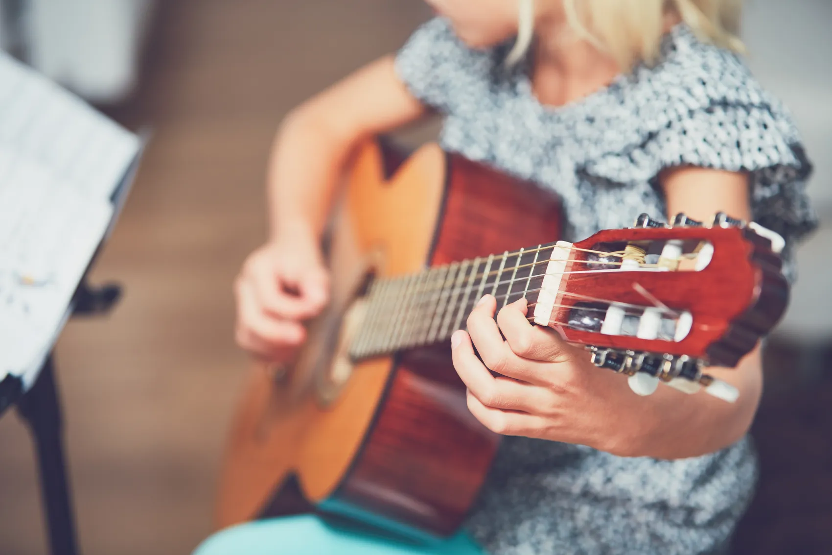 Kind spielt Gitarre, die Finger greifen die Saiten. Notenblätter stehen auf einem Notenständer im Hintergrund.
