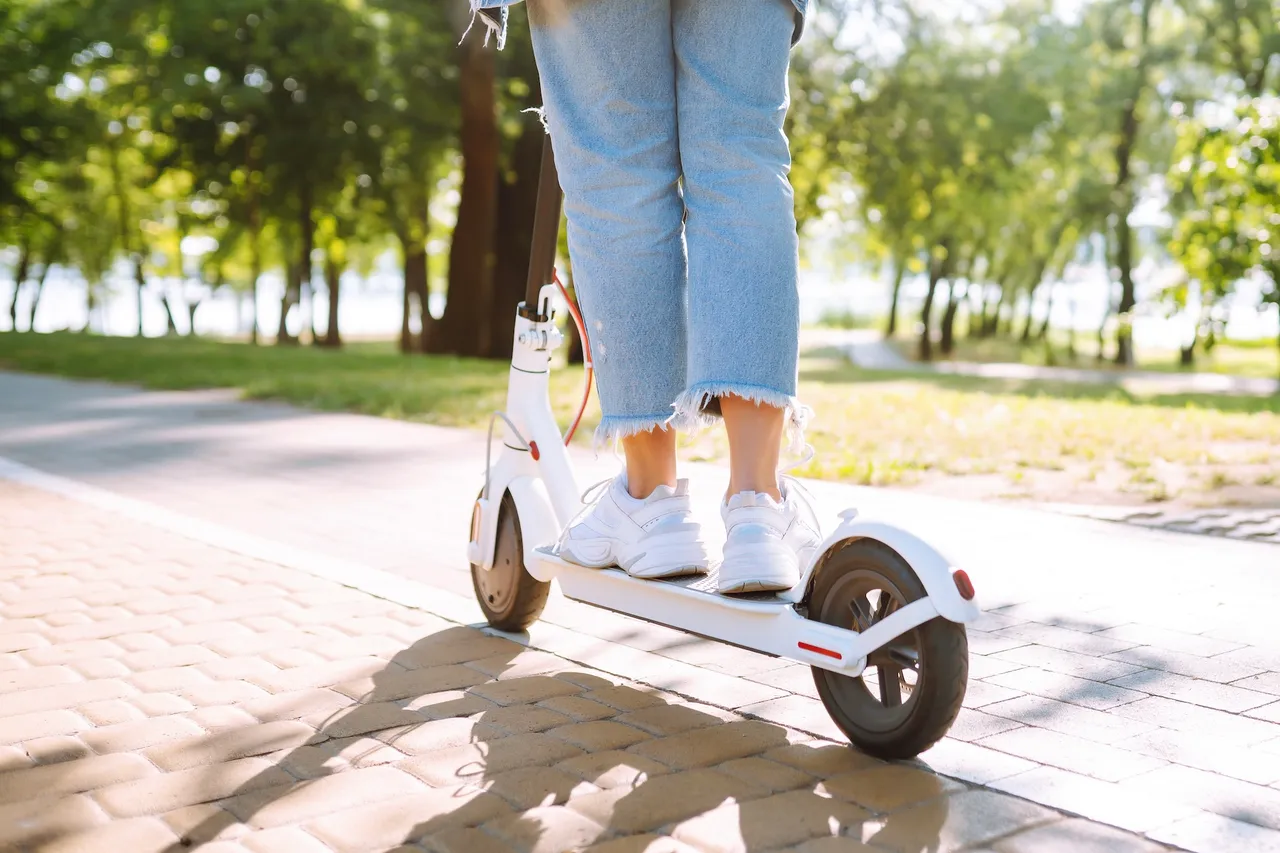 Eine Person fährt auf einem weißen E-Scooter auf einem gepflasterten Weg durch einen Park.