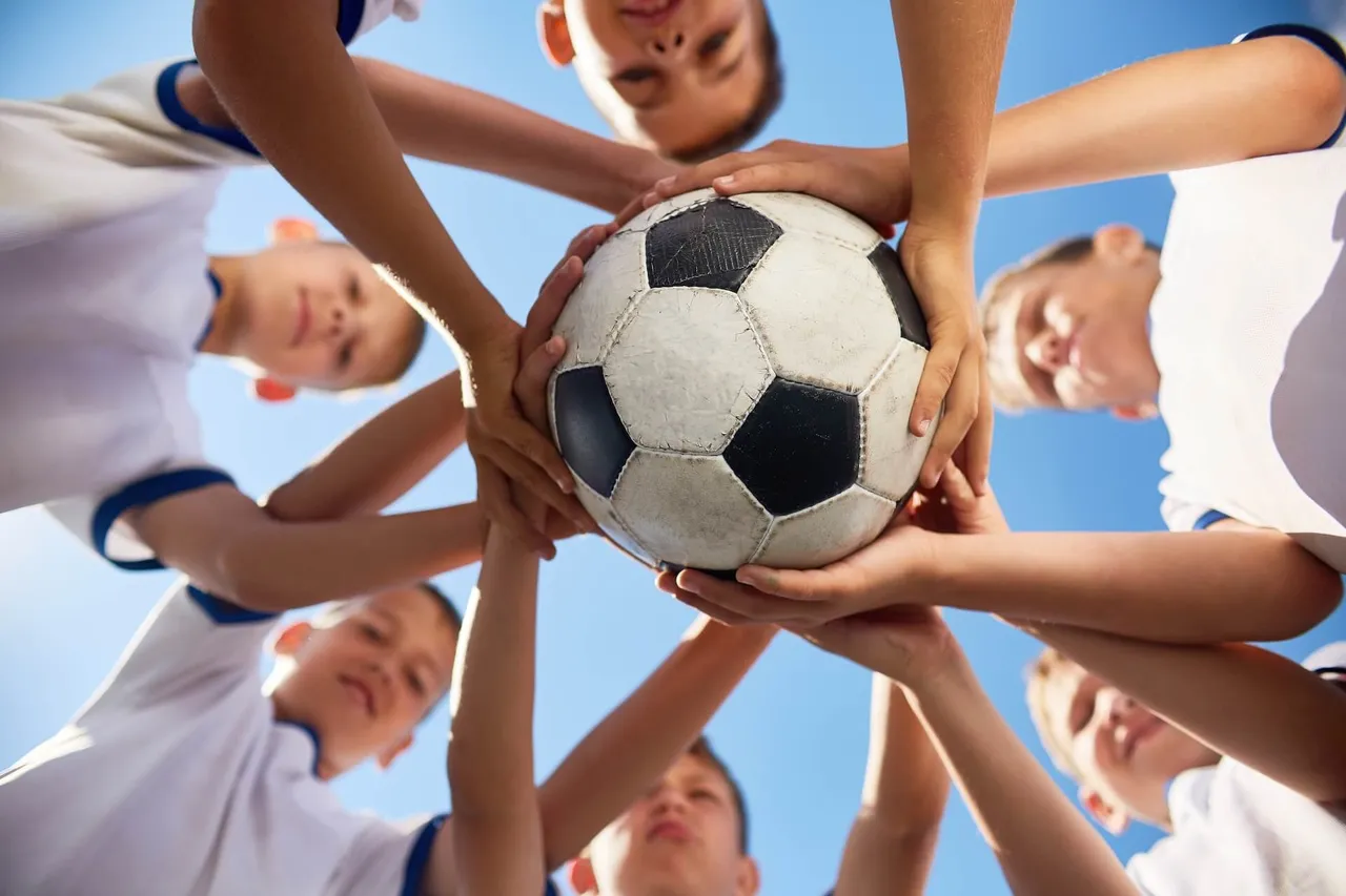 Eine Gruppe Jugendlicher in weißen Fußballtrikots umringt einen Fußball und legt ihre Hände darauf.